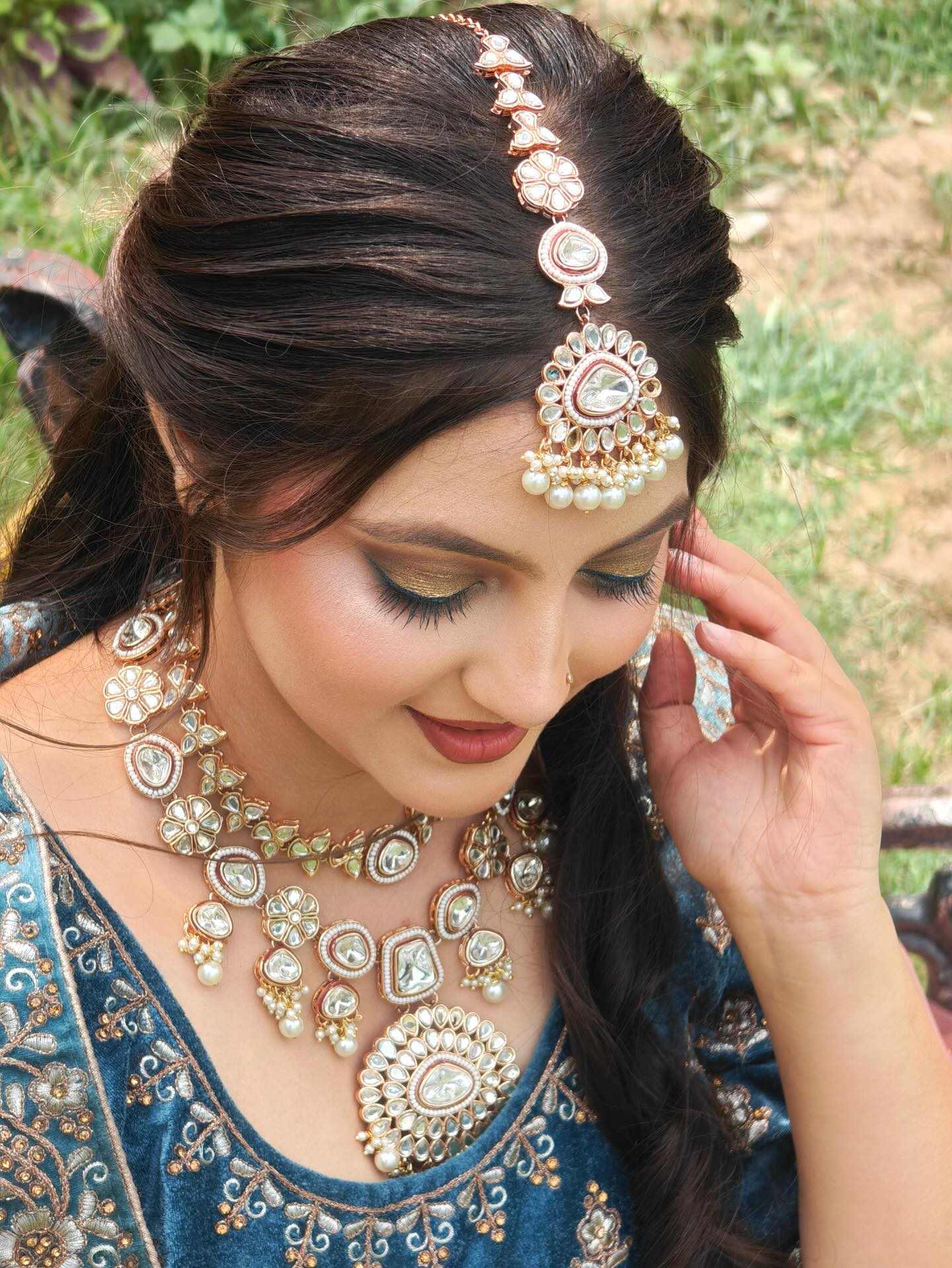 Woman with long dark hair styled in soft waves, wearing an ornate blue and gold embroidered outfit and elaborate gold and pearl jewelry including a necklace and headpiece, sitting outdoors on a bench with green grass and plants in the background. The setting feels festive and elegant, suggesting a special occasion or celebration.