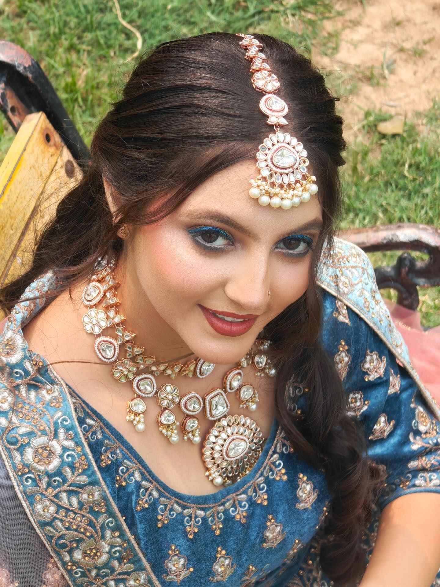 Woman wearing a blue and gold embroidered traditional outfit, adorned with ornate gold and pearl jewelry including a necklace and headpiece, sitting outdoors on a bench with green grass in the background. The scene conveys a festive and elegant atmosphere.