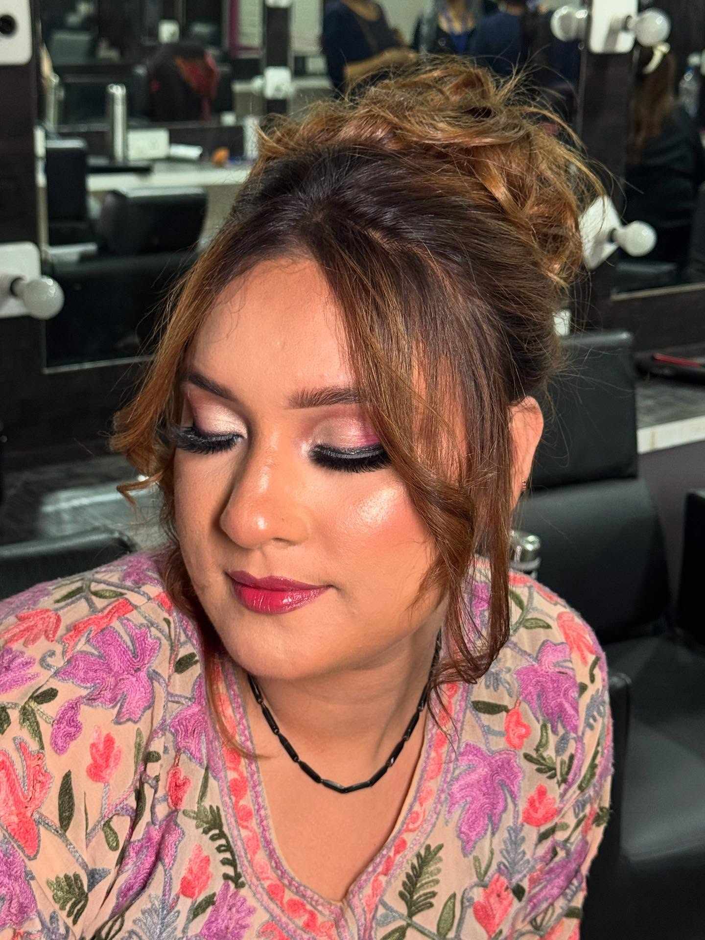 Woman with styled brown hair wearing a floral embroidered top and a black beaded necklace, seated in a modern salon with black leather chairs and mirrors in the background. The environment is professional and welcoming, suggesting a beauty or makeover session.