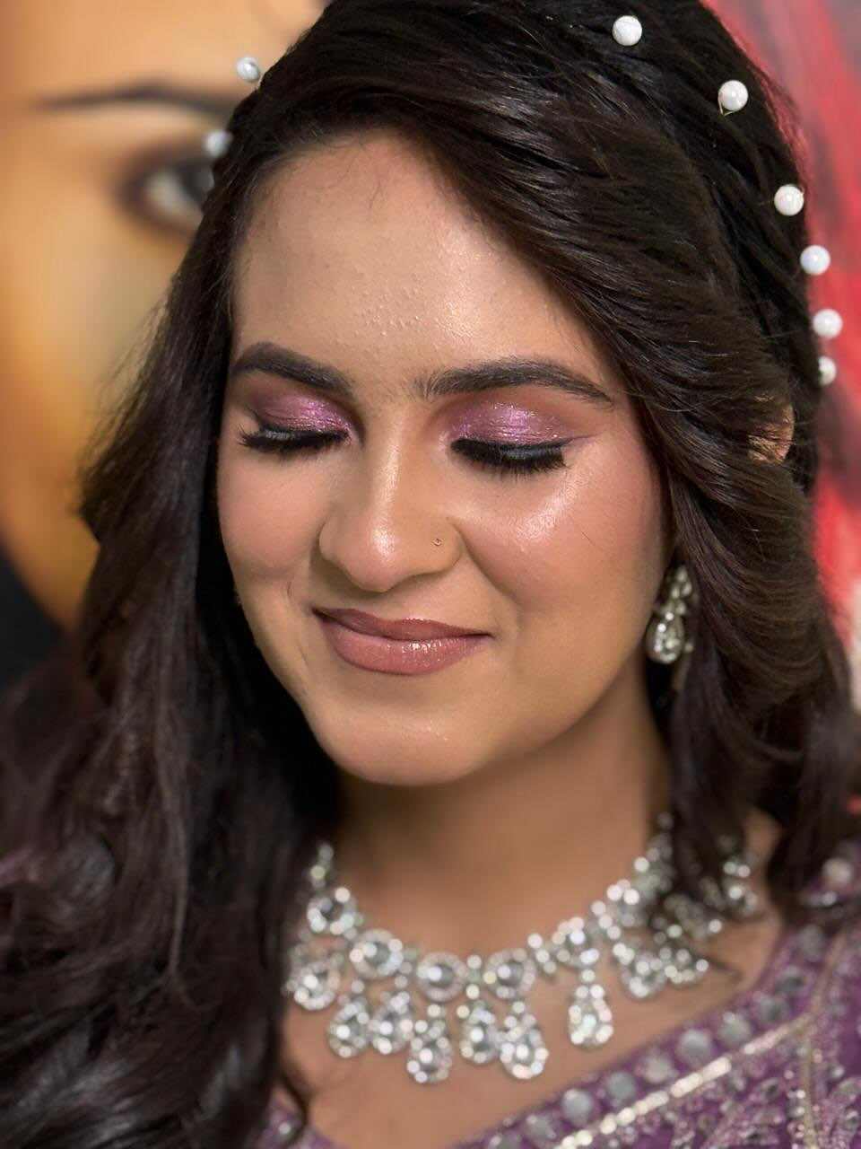 Woman with long wavy dark hair adorned with pearl hairpins, wearing a sparkling silver necklace and matching earrings, dressed in a purple and silver embroidered outfit. The background features warm colors and a festive atmosphere, suggesting a celebratory or special occasion. The overall tone is elegant and joyful.